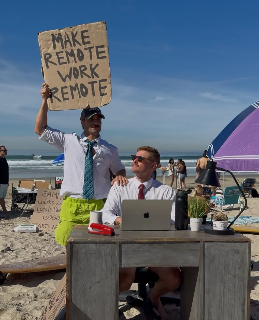 Two men at a beach, one seated at a desk with a laptop and the other holding a sign that reads "MAKE REMOTE WORK REMOTE."