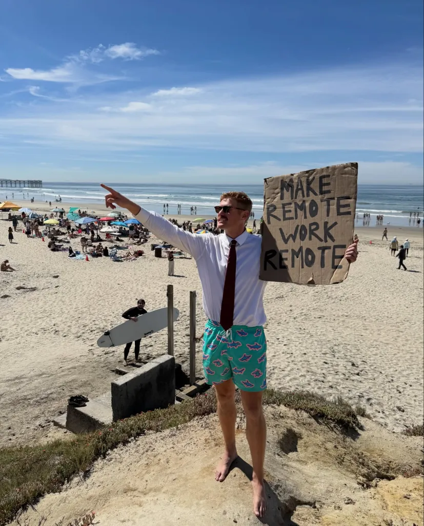 Scott Muirhead on a beach holding a sign that says "Make Remote Work Remote".
