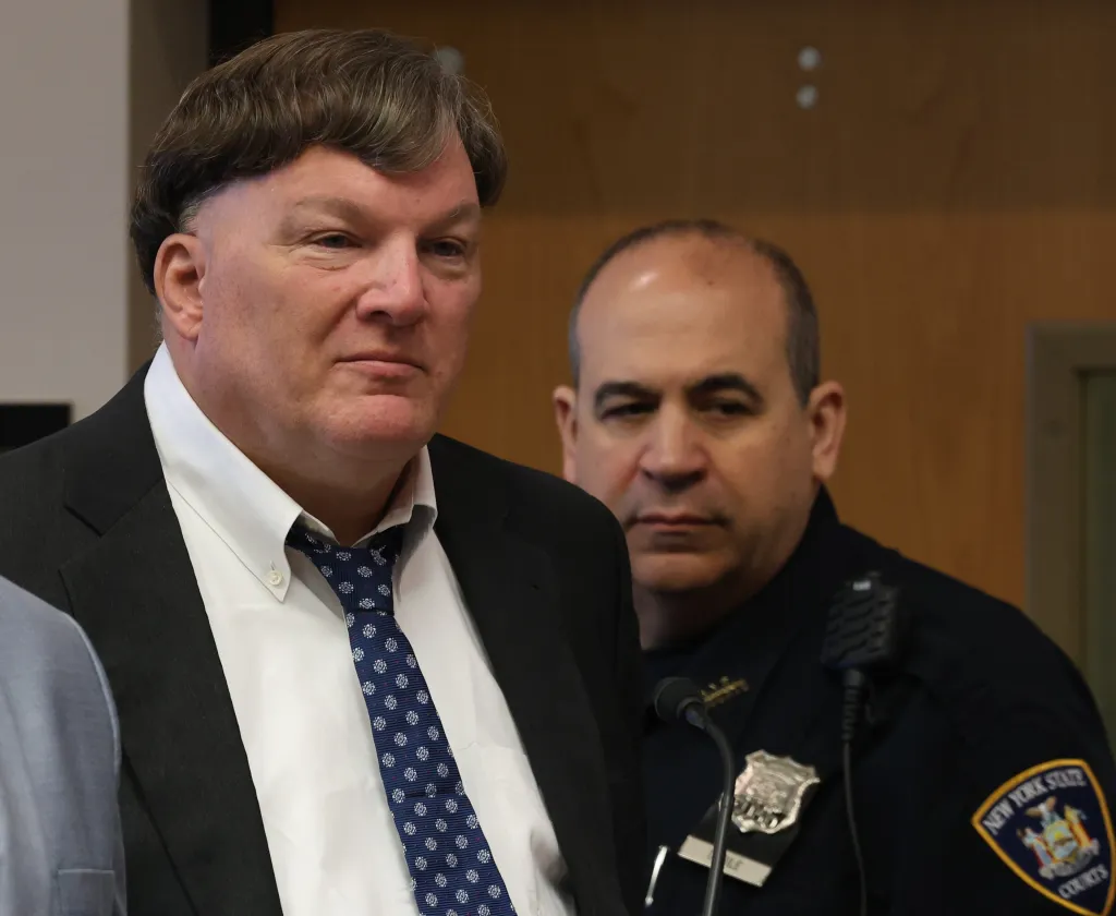 Rex Heuermann, the Gilgo Beach serial killer, is shown in court, with a police officer standing behind him.