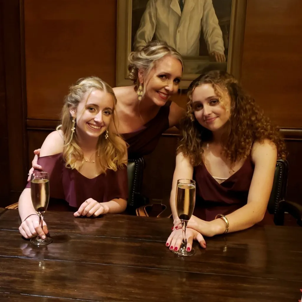 Amy Hillyard with two young women, all wearing dark red dresses, smiling and holding champagne flutes at a wooden table.