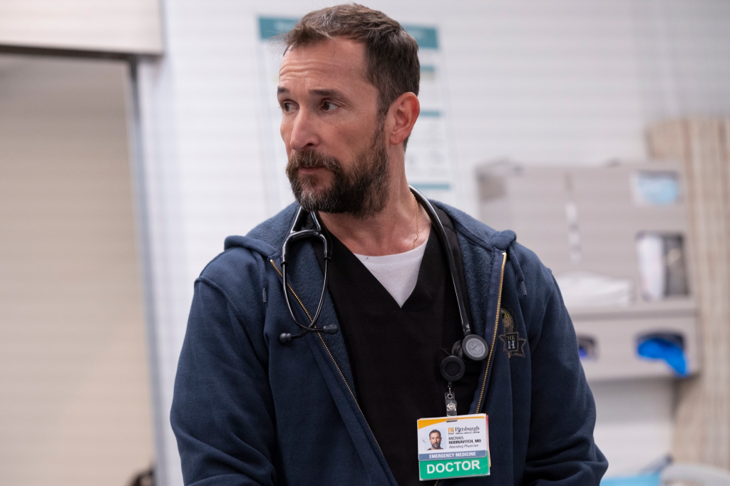 A male doctor with a beard wearing a dark blue zip-up hoodie over scrubs, with a stethoscope around his neck and a "Doctor" ID badge, looking to the side.