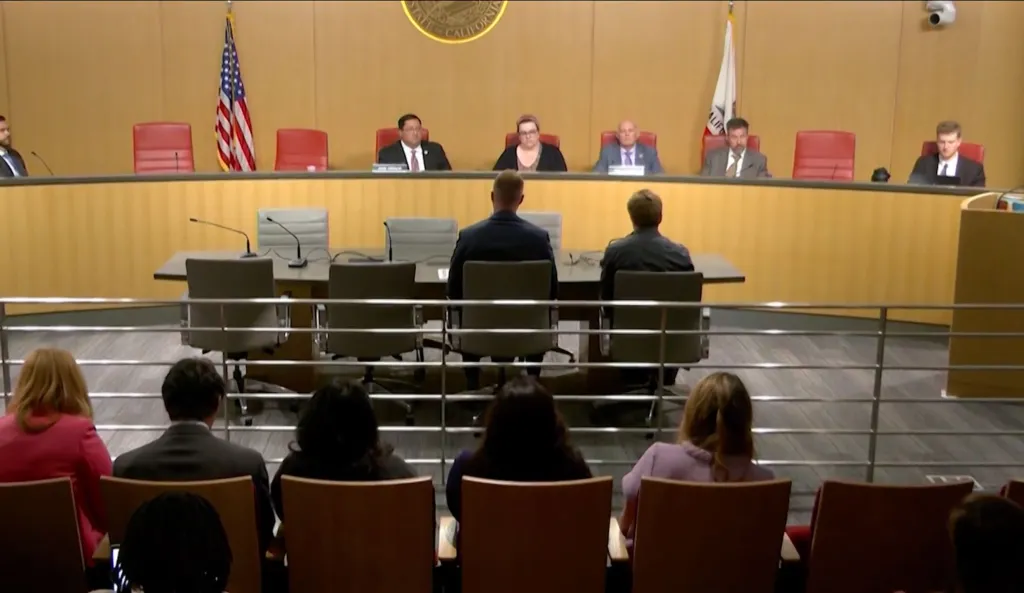 Robert Scrivner, son, sitting at a table with other men and women, with an American flag and a Californian flag in the background.