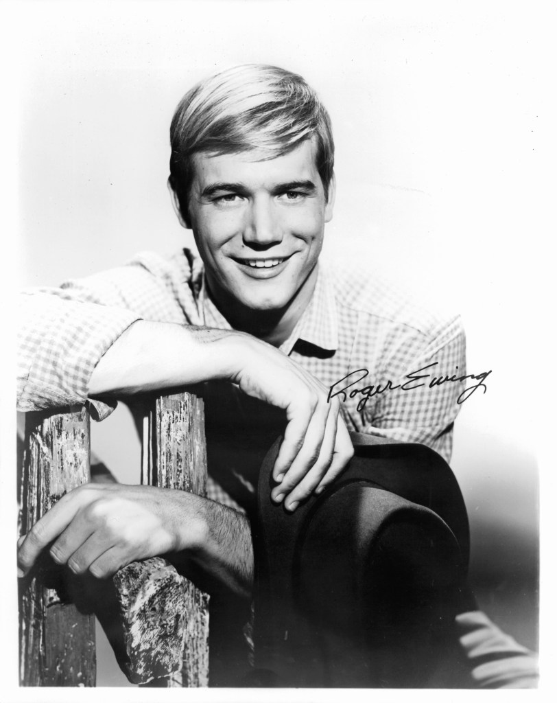 Roger Ewing smiling, leaning on a wooden fence, holding a hat.