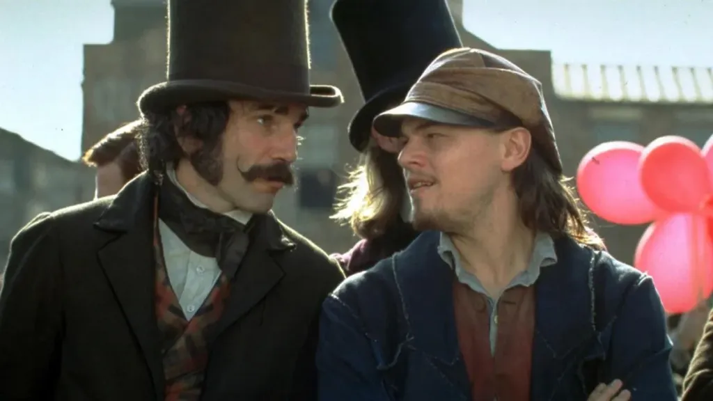Two men in 19th-century attire stand close together in conversation. The man on the left wears a tall top hat, a dark coat, and sports thick mutton chop sideburns and a curled mustache. The man on the right wears a brown cap and rugged clothes with a slightly scruffy beard. Behind them, a bunch of pink balloons adds contrast to the otherwise muted historical setting.