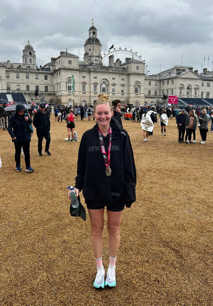 Molly Baker smiling, wearing a medal, black hoodie, and holding a water bottle, with Horse Guards Parade and the London Eye in the background.