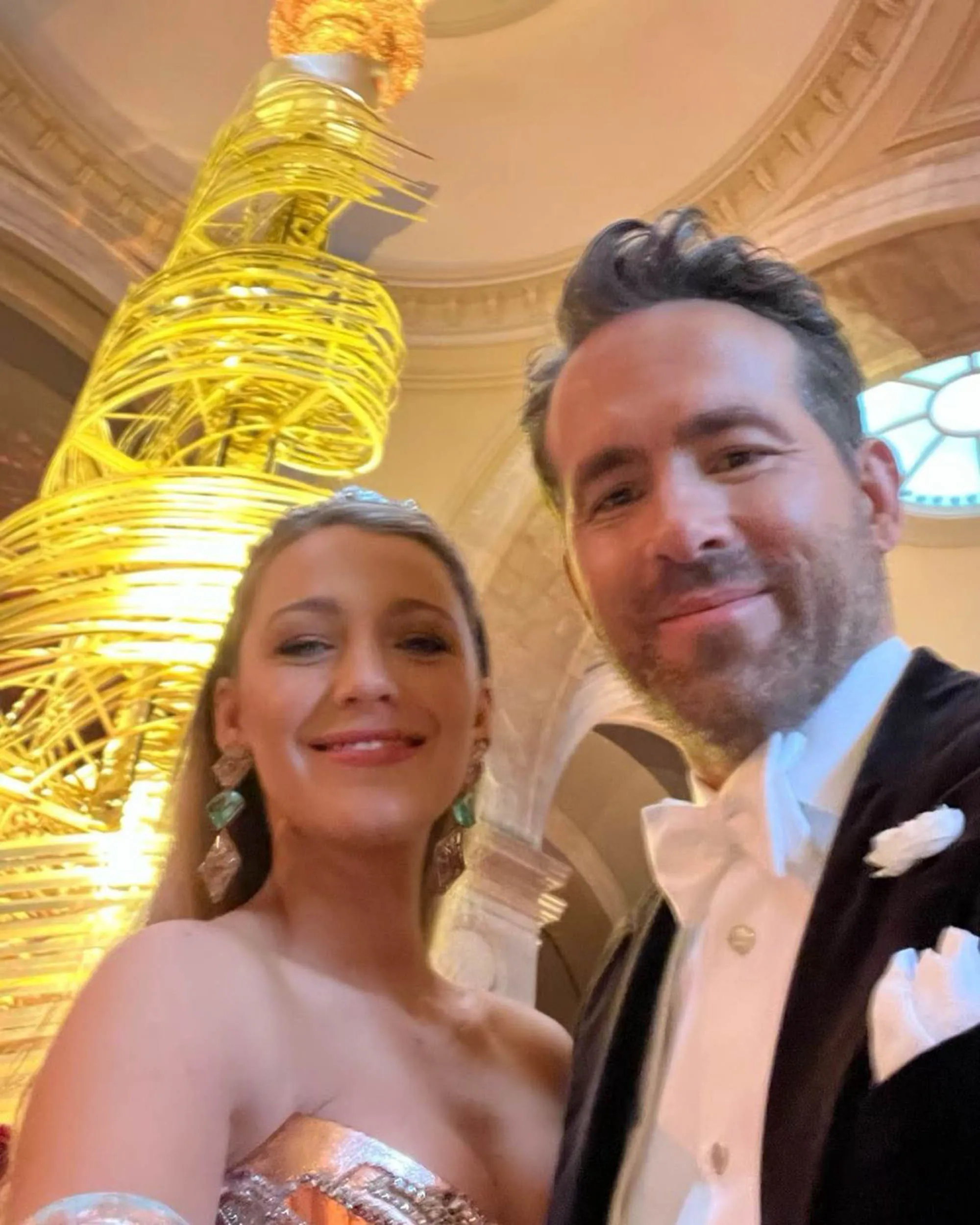 Ryan Reynolds and Blake Lively smiling at the camera, with a large golden structure behind them.