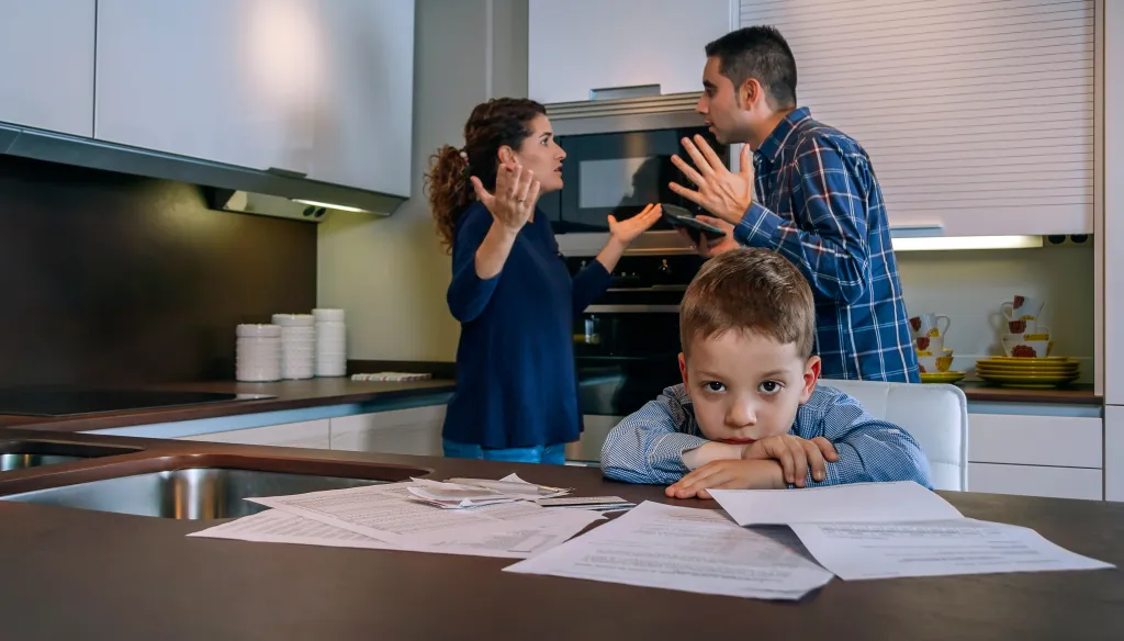 A sad boy looks at the camera while his parents argue in the background of their kitchen.