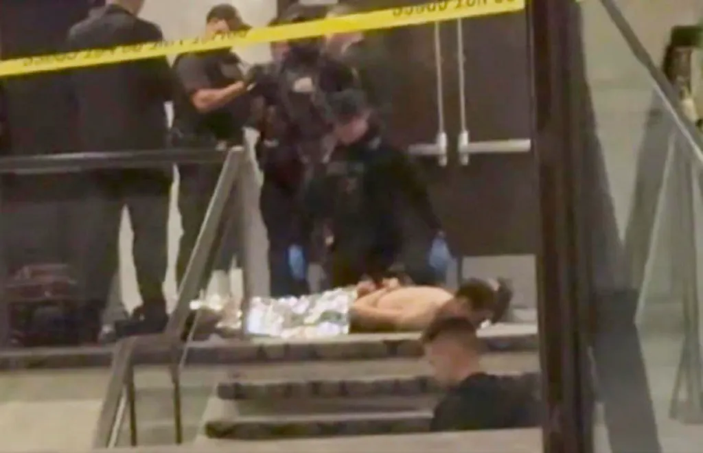 Law enforcement officers surround a shirtless individual lying on stairs under a yellow "DO NOT CROSS" tape.