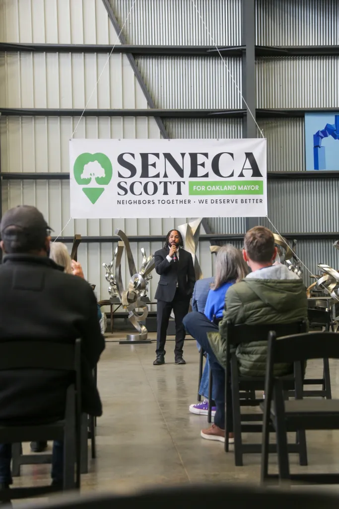 Seneca Scott speaking at an event under a banner that reads "Seneca Scott for Oakland Mayor, Neighbors Together, We Deserve Better".
