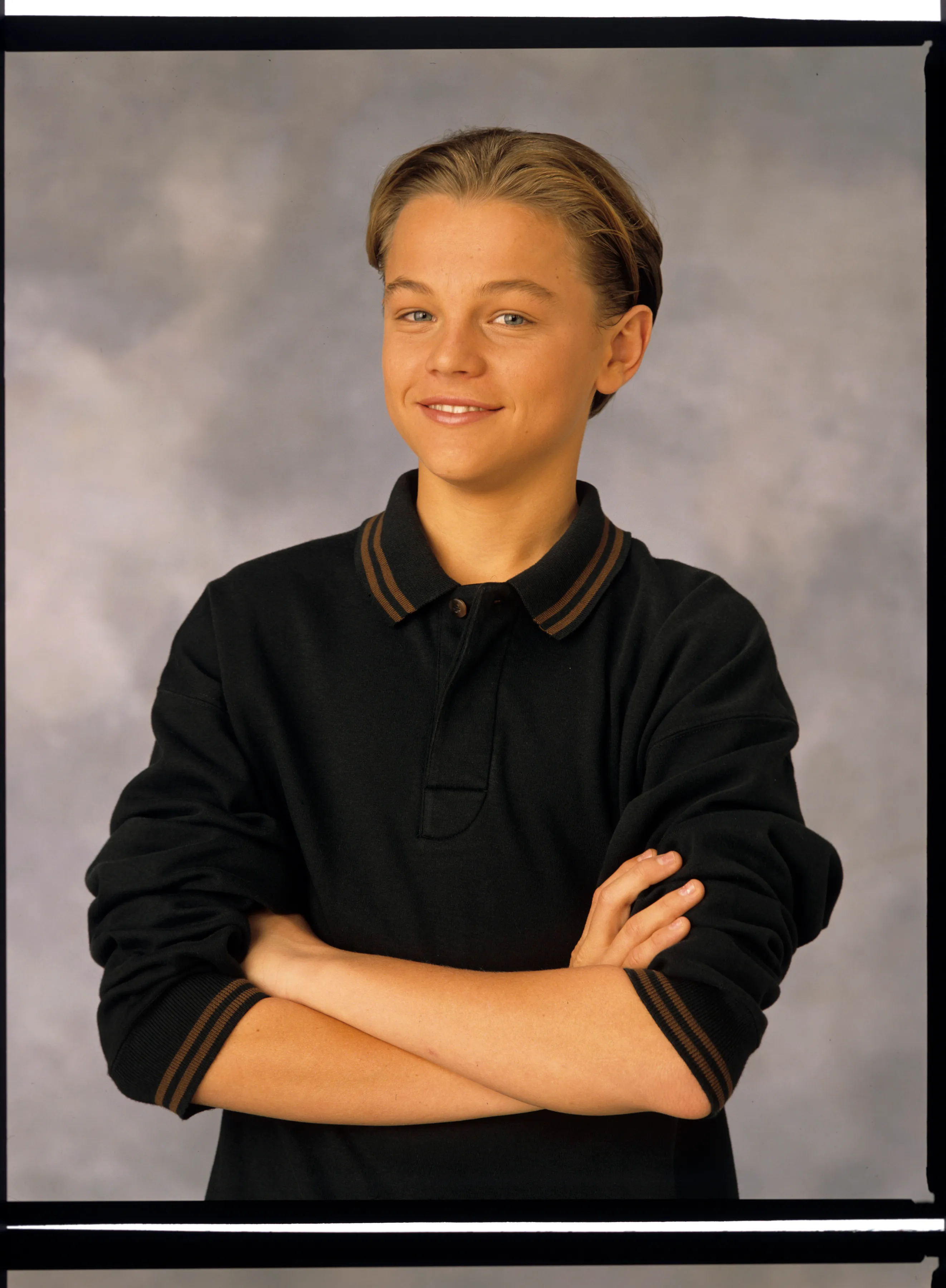 Young Leonardo DiCaprio in a black polo shirt with brown stripes on the collar and sleeves, smiling with his arms crossed.