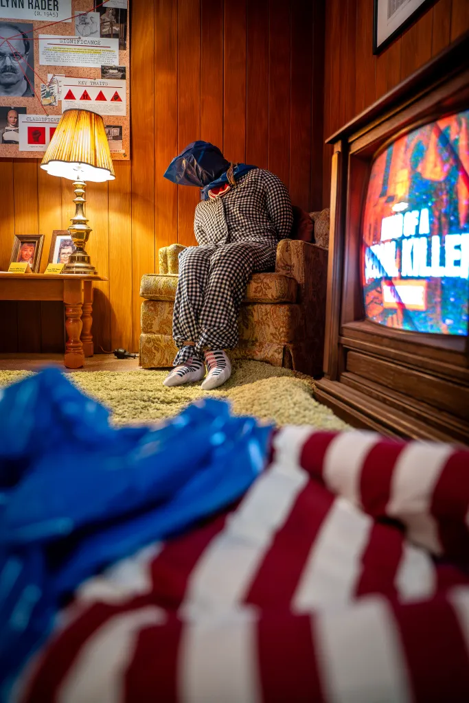 An exhibit features a mannequin with a blue bag over its head and hands bound, seated in a patterned armchair, with a TV in the foreground displaying "A Serial Killer" and information about Dennis Rader (BTK killer) on the wall.
