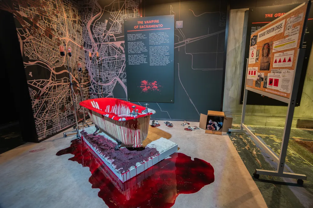 A bloody bathtub sits center in a room with a large map on the back wall and a corkboard display to the right, all part of a "Serial Killer Exhibit NYC."
