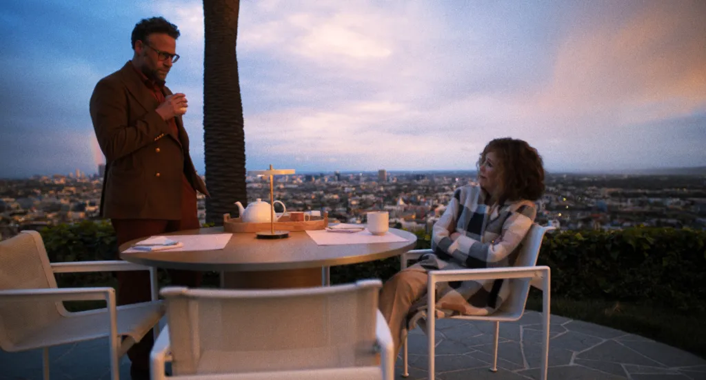 Seth Rogen and Catherine O'Hara sitting face to face at a table outdoors overlooking a city at dusk.