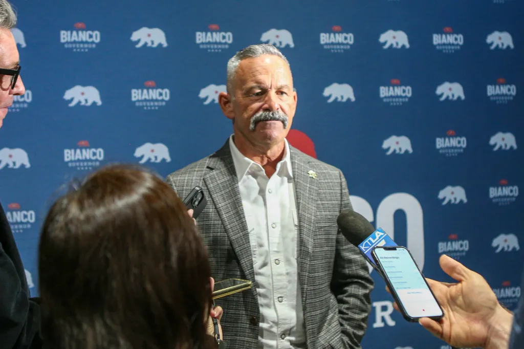 Sheriff Chad Bianco speaks at a press conference with "BIANCO GOVERNOR" signs in the background.