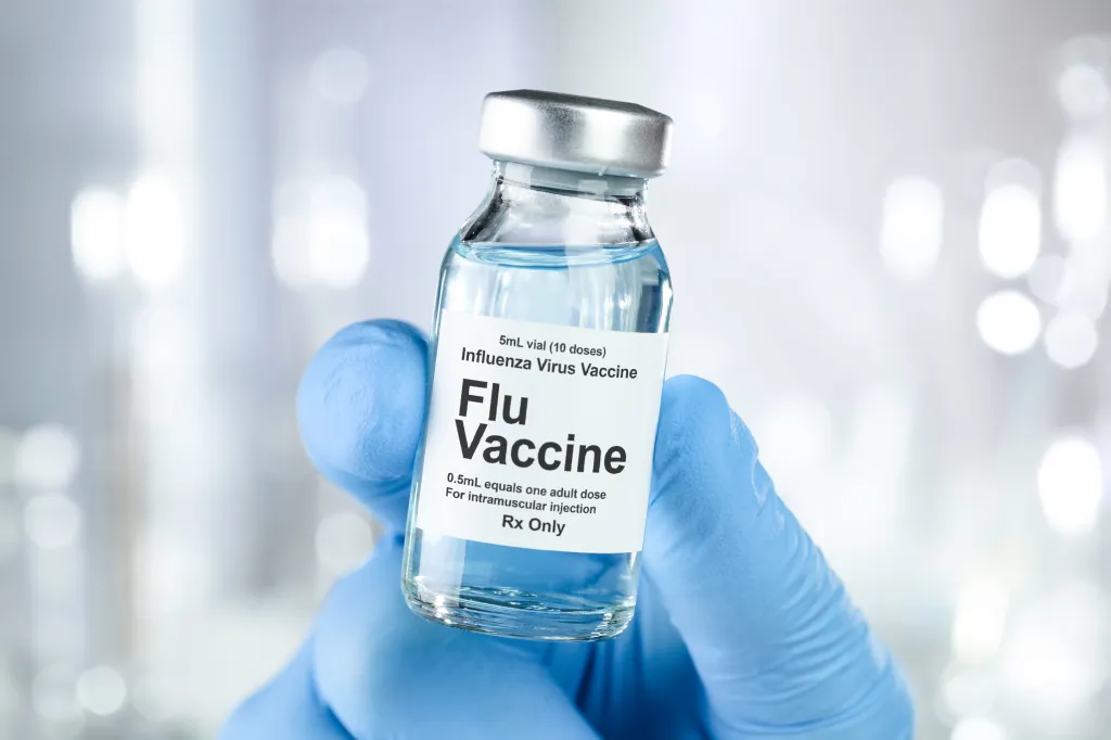 A gloved hand holds a vial labeled "Flu Vaccine" for influenza.