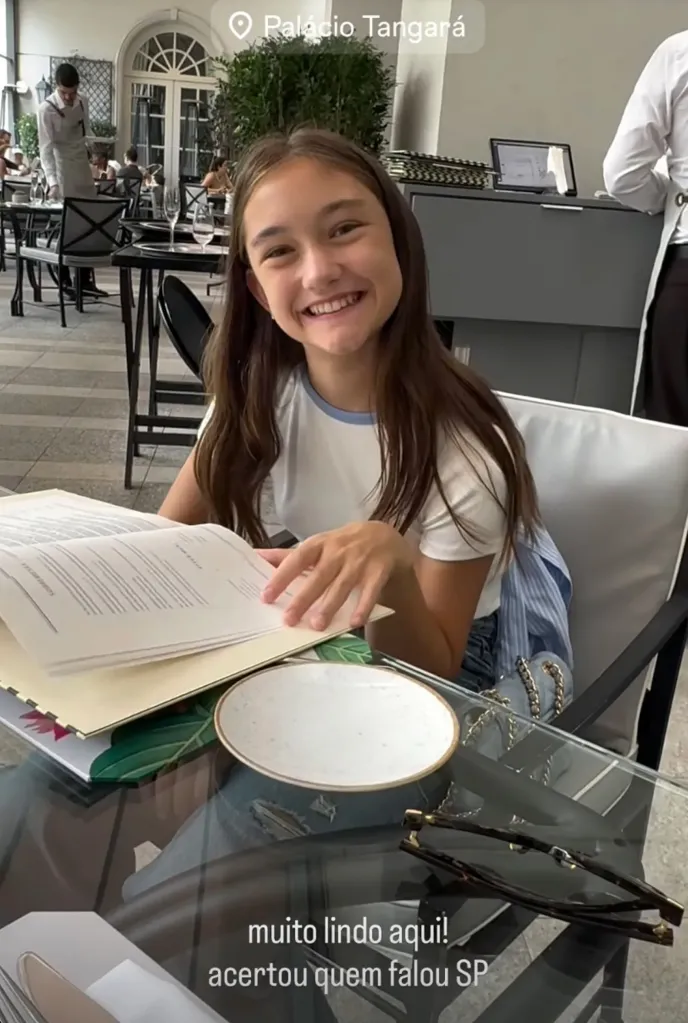 A young girl sits at a table in a restaurant, looking up and smiling.