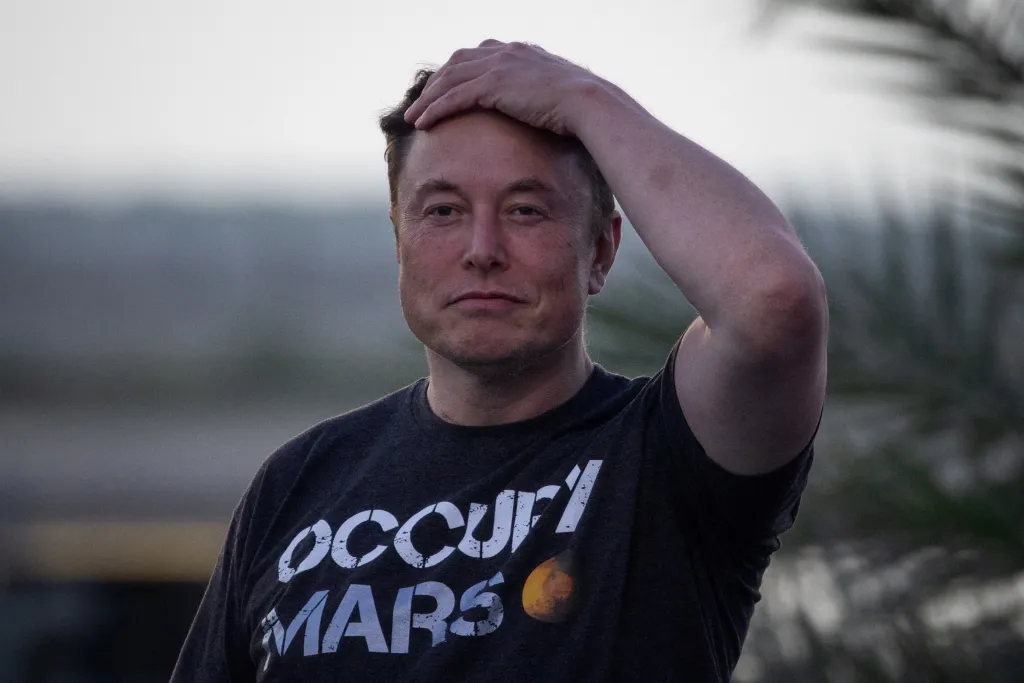 Elon Musk wearing an "Occupy Mars" t-shirt, with one hand on his head.