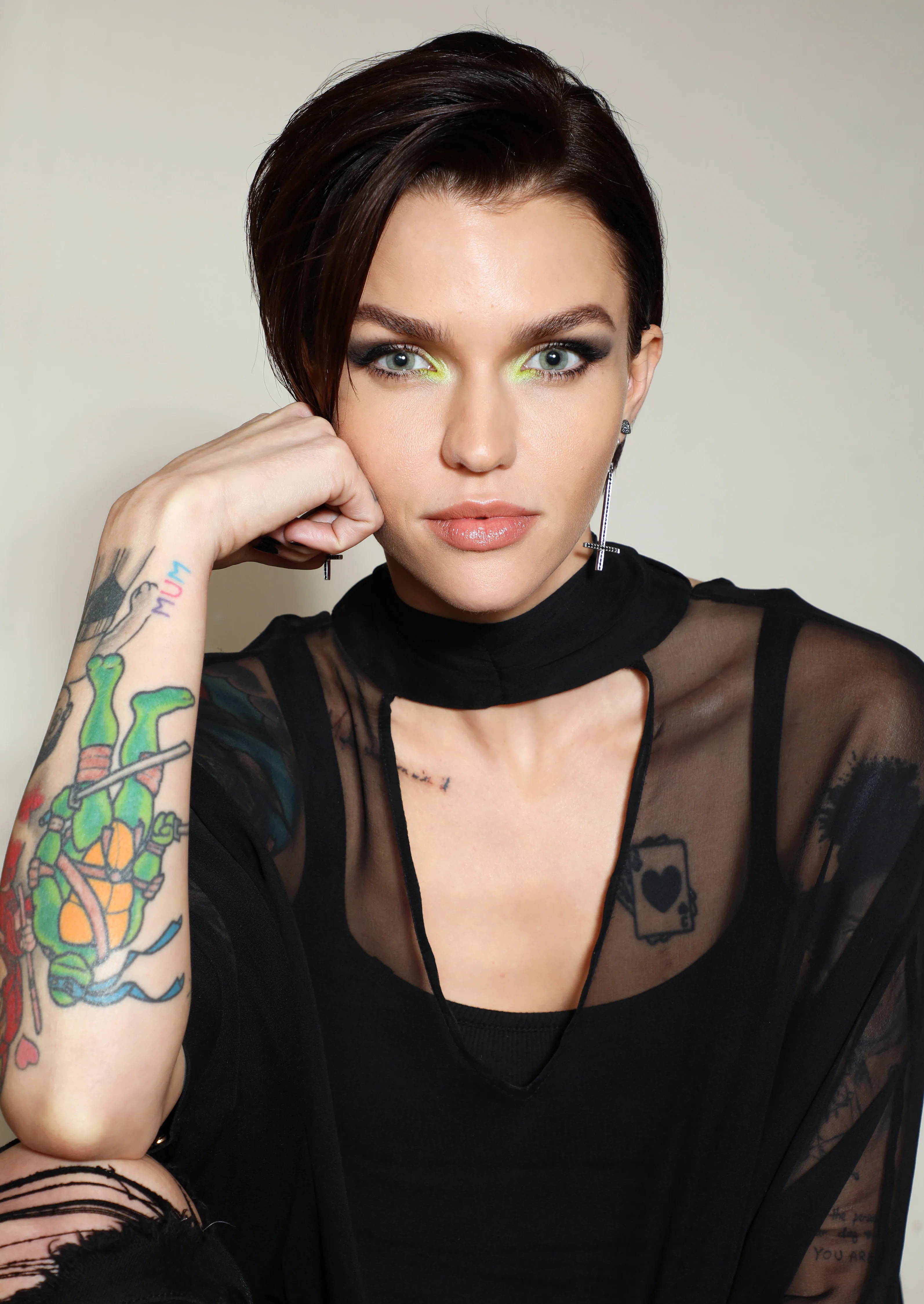 Ruby Rose posing with her hand to her chin, wearing a sheer black top and cross earrings.