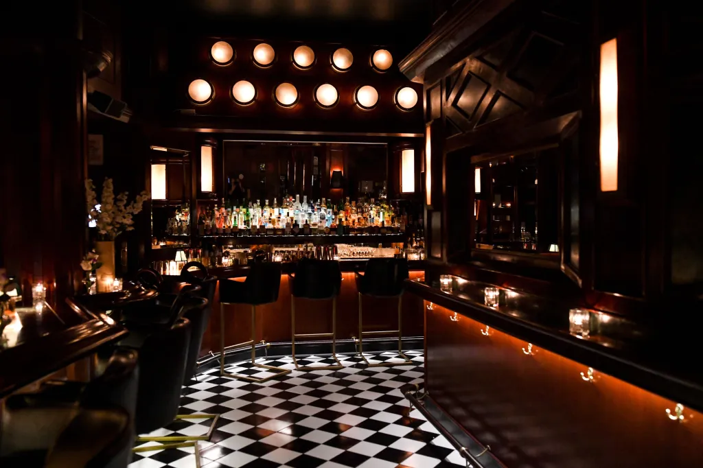 A dimly lit bar with a checkerboard floor, a long bar with bottles of alcohol, and bar stools.