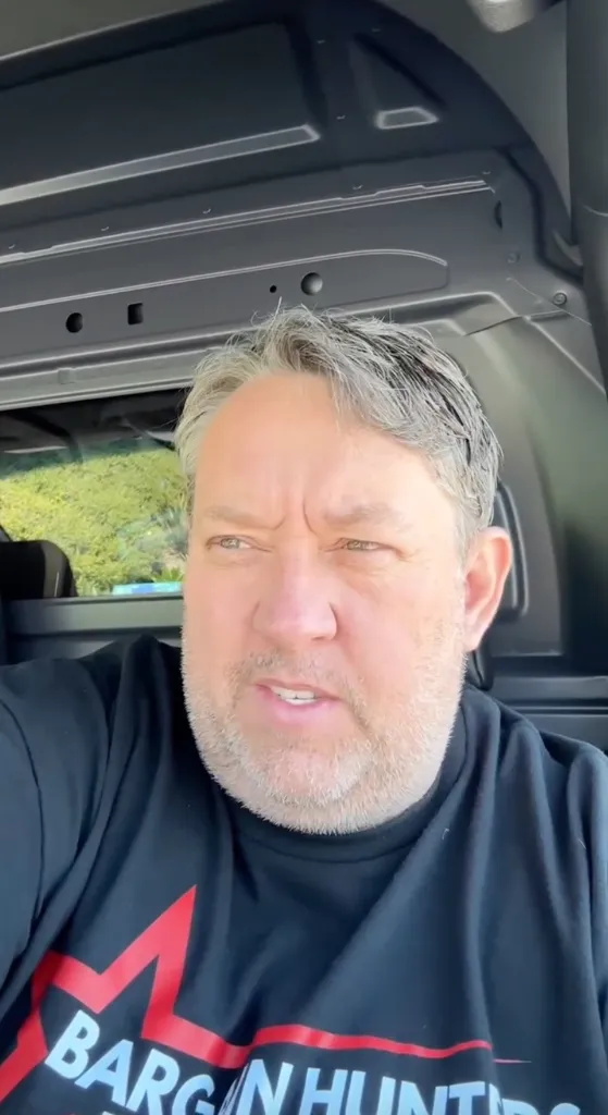 A man with graying hair and a beard, wearing a black t-shirt with "BARGAIN HUNTERS" on it, sits in a vehicle.