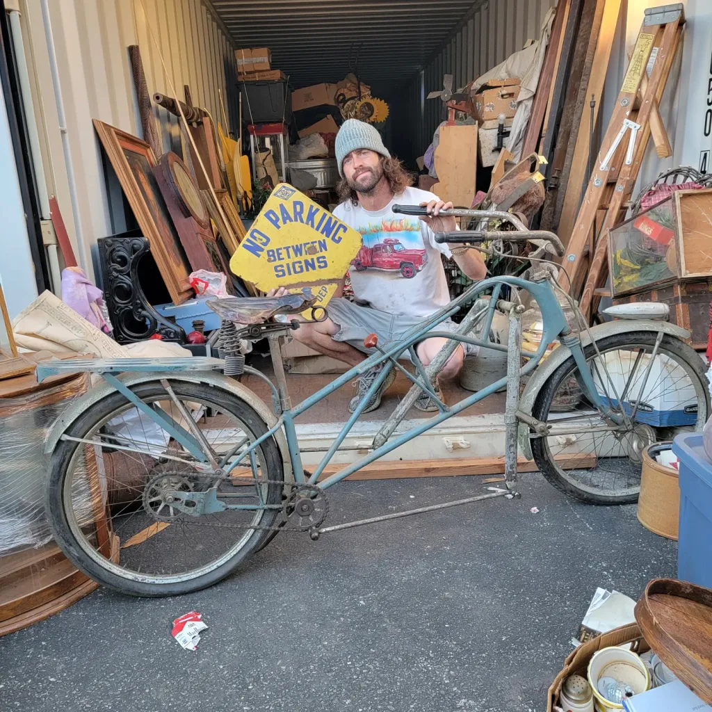 Man holding a "NO PARKING BETWEEN SIGNS" sign, next to a vintage bicycle in a storage unit.