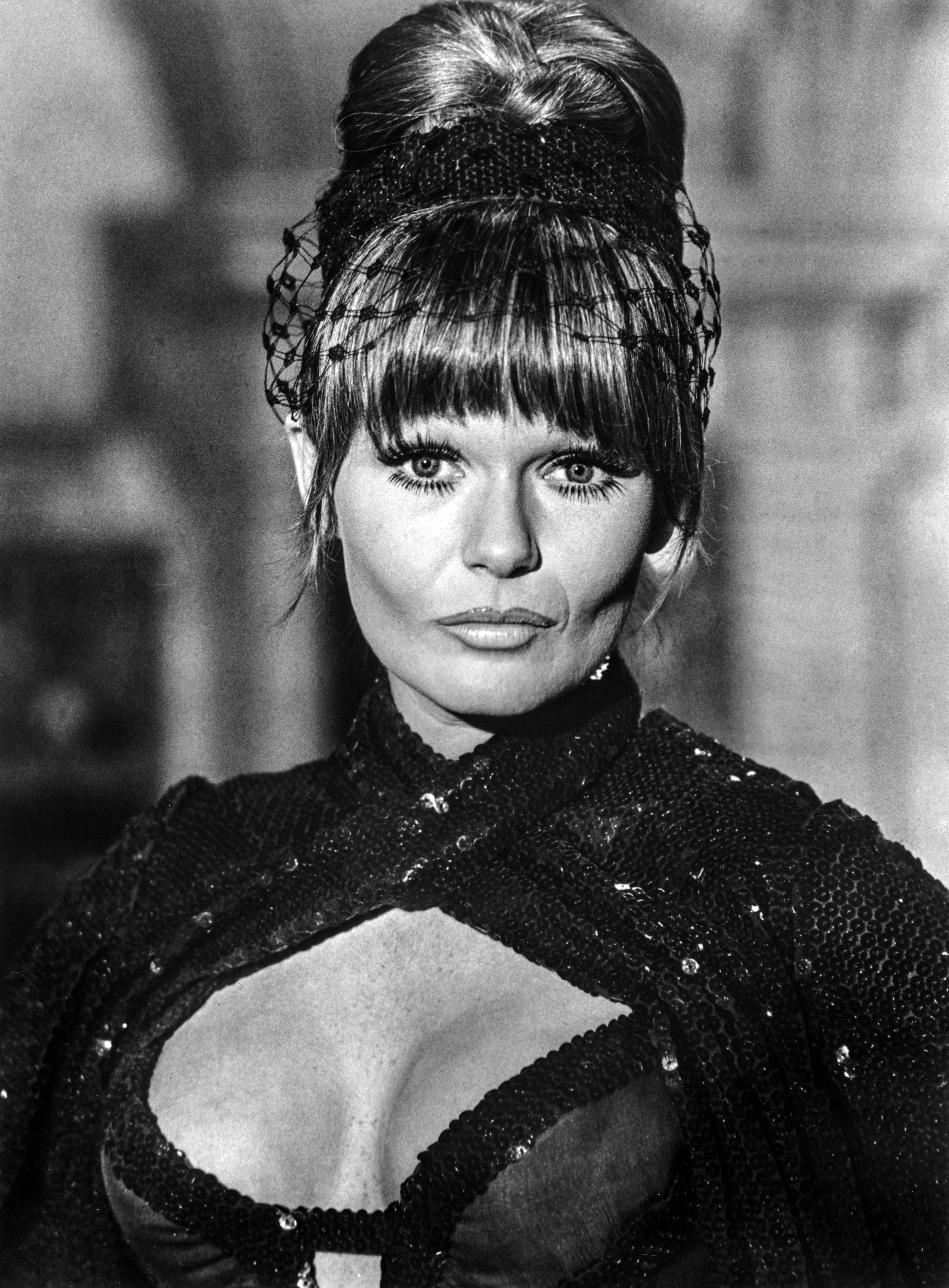Portrait of a woman with an elaborate updo and a net veil over her bangs, wearing a sequined top with a plunging neckline.