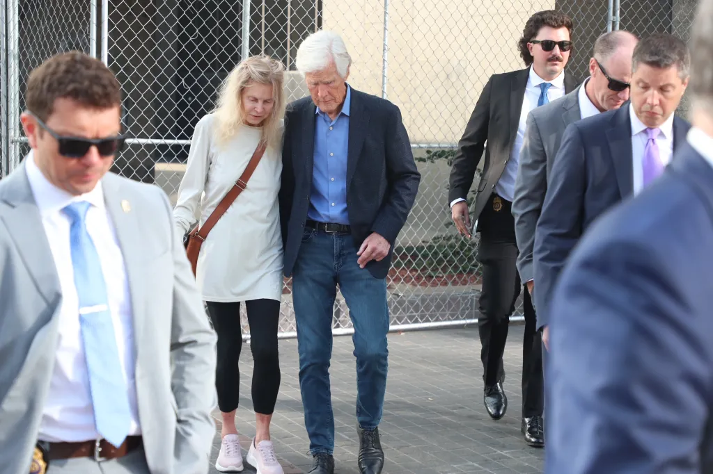 Suzanne Perry and Keith Morrison walking with other men in suits, with a chain link fence behind them.