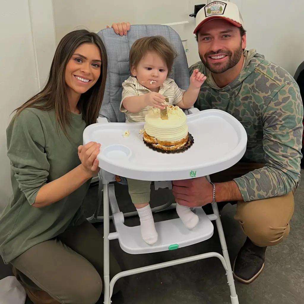 A woman, man, and baby celebrate a first birthday with a small cake on a highchair.