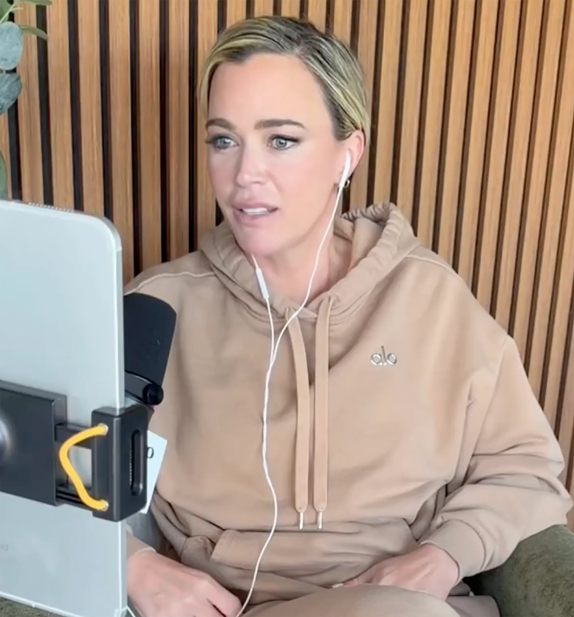 Teddi Mellencamp in a beige Alo Yoga hoodie, wearing wired earbuds and looking at a tablet in a stand.