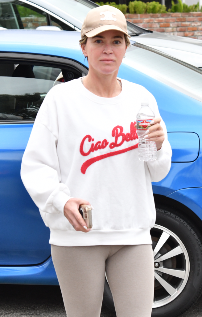 Teddi Mellencamp in a white "Ciao Bella" sweatshirt, tan leggings, and a baseball cap, holding a water bottle and phone.