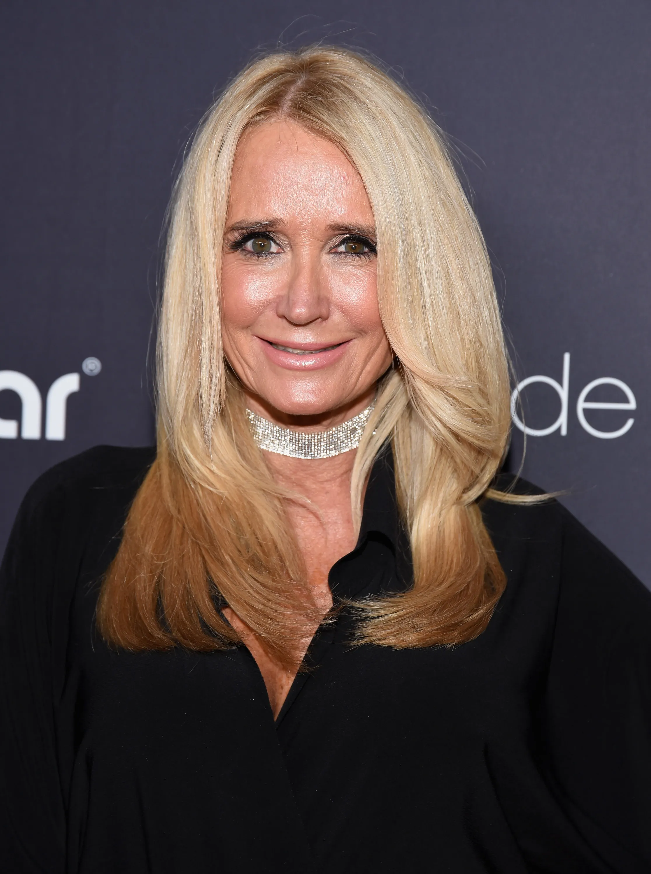 Kim Richards, a television personality, smiles at the camera while wearing a sparkly choker and a black top.