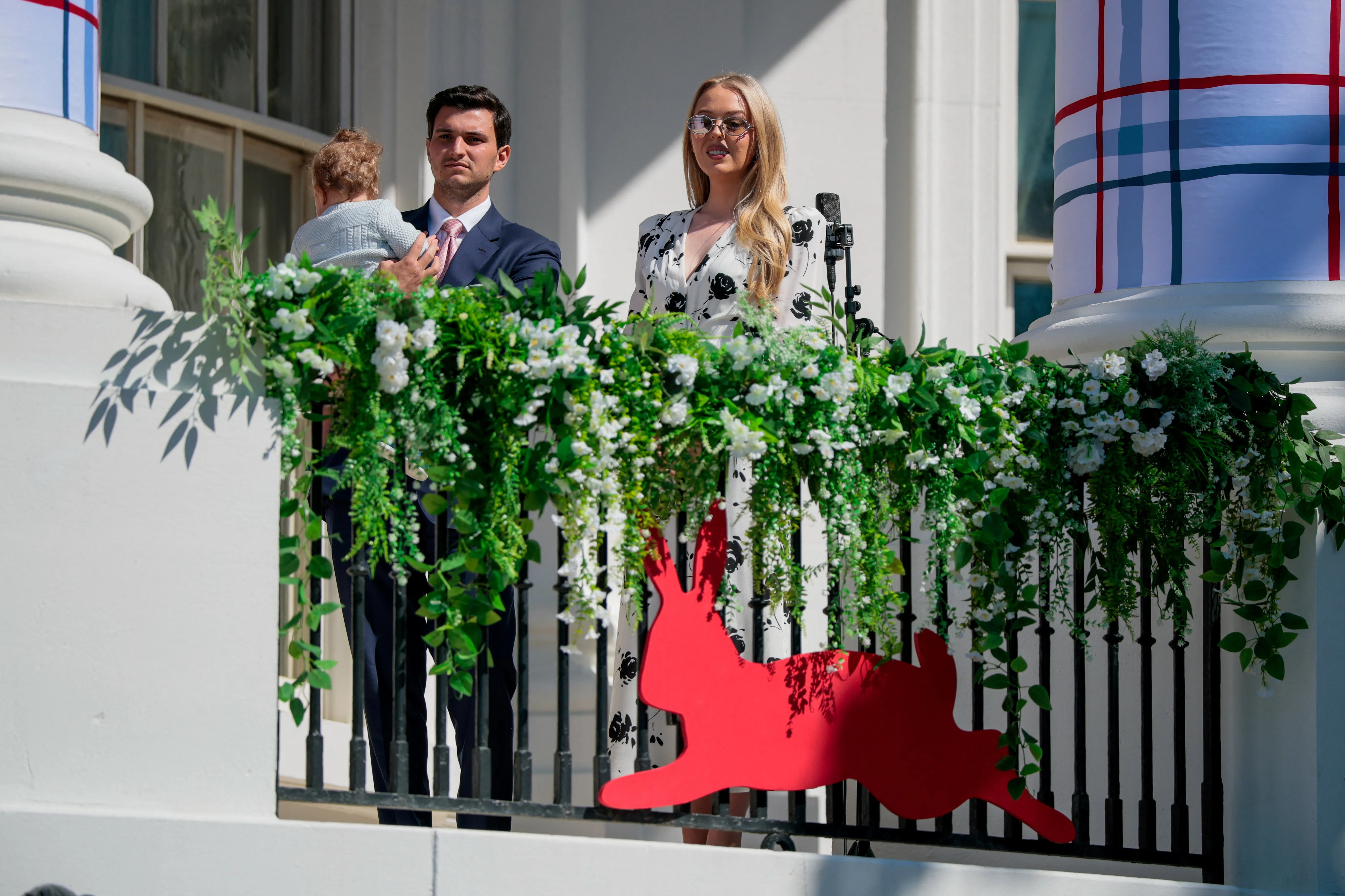 Tiffany Trump and Michael Boulos stand on a balcony, holding a baby, with an Easter rabbit decoration and floral garlands.