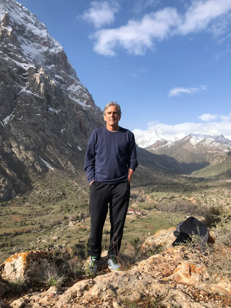 Barry Hoffner standing on a rocky outcrop in Tajikistan, with snow-capped mountains and a valley behind him.