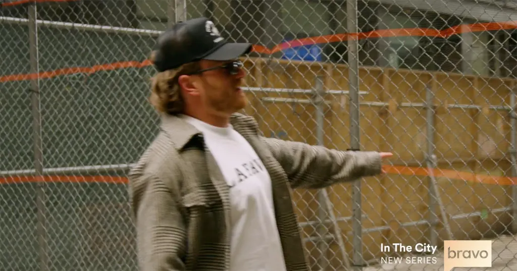 A man in a black cap and sunglasses points to the right, standing in front of a chain-link fence.