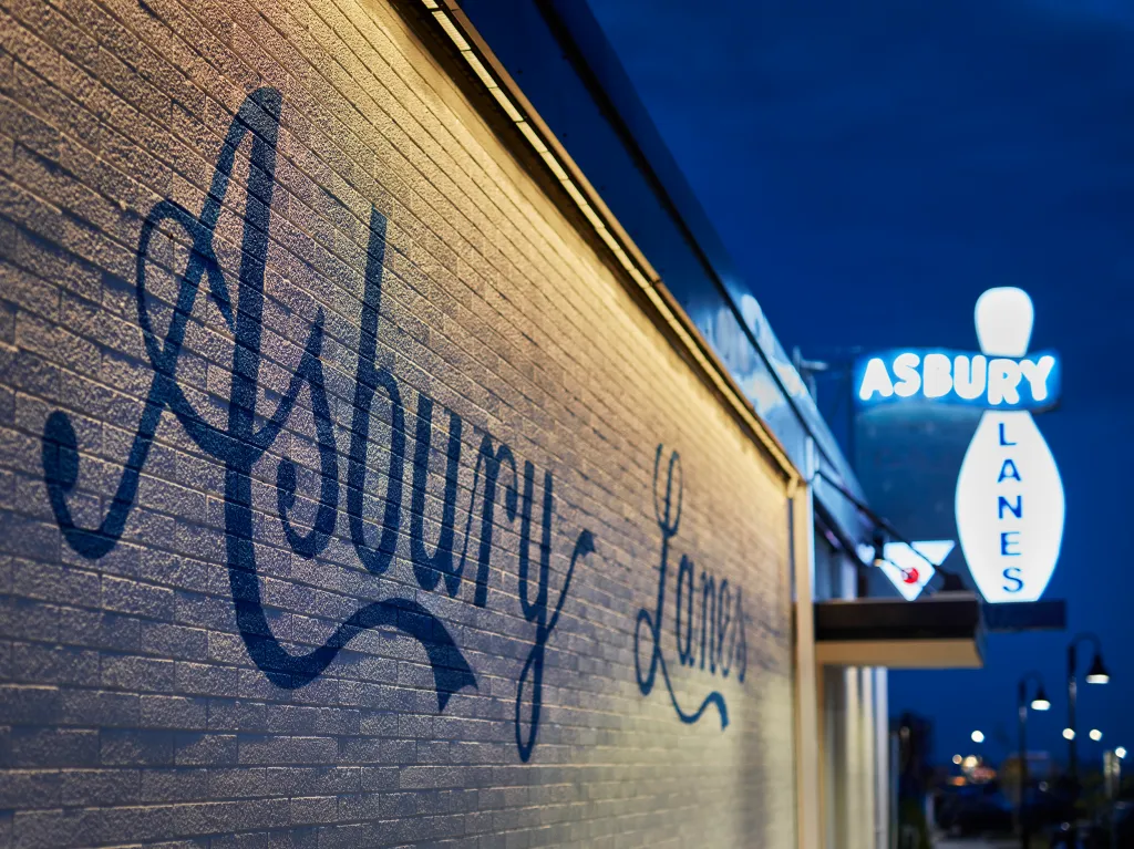 The exterior of Asbury Lanes at the Asbury Hotel at night, with "Asbury Lanes" in blue script on a brick wall and a glowing neon bowling pin sign.