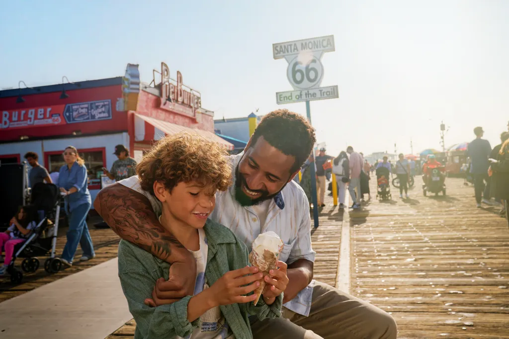 A man and child on the Santa Monica Pier, in front of the Route 66 "End of the Trail" sign, sharing ice cream.