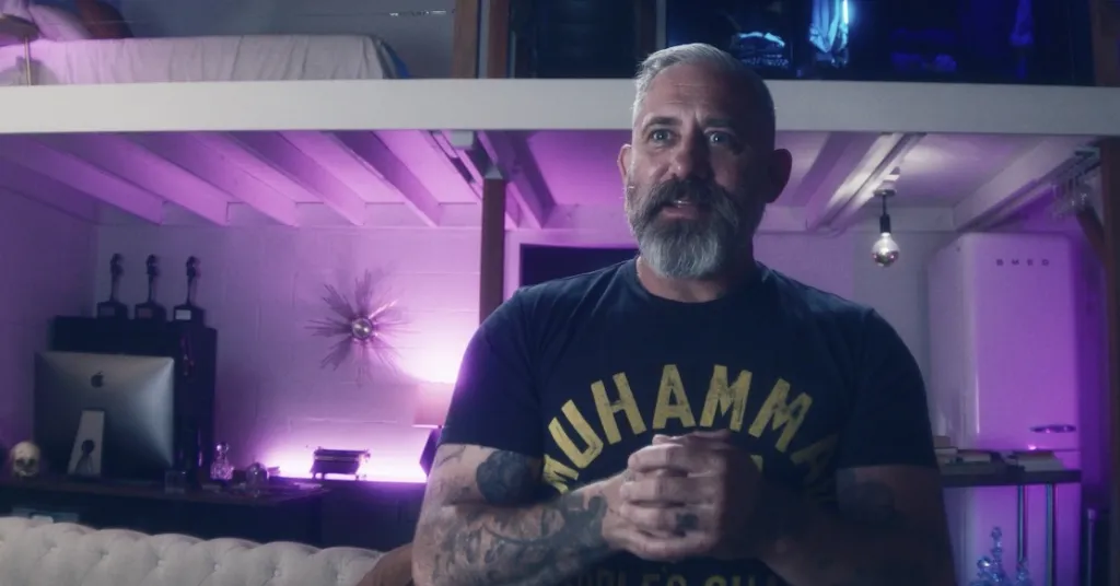 A man with a beard, tattoos, and a "MUHAMMAD" T-shirt stands in a room with purple lighting.