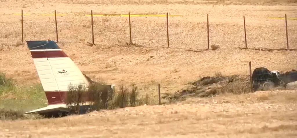 The tail section of a crashed single-engine Piper airplane, with "Piper" visible on the tail, lies on a dry, grassy field next to a fence with yellow police tape.