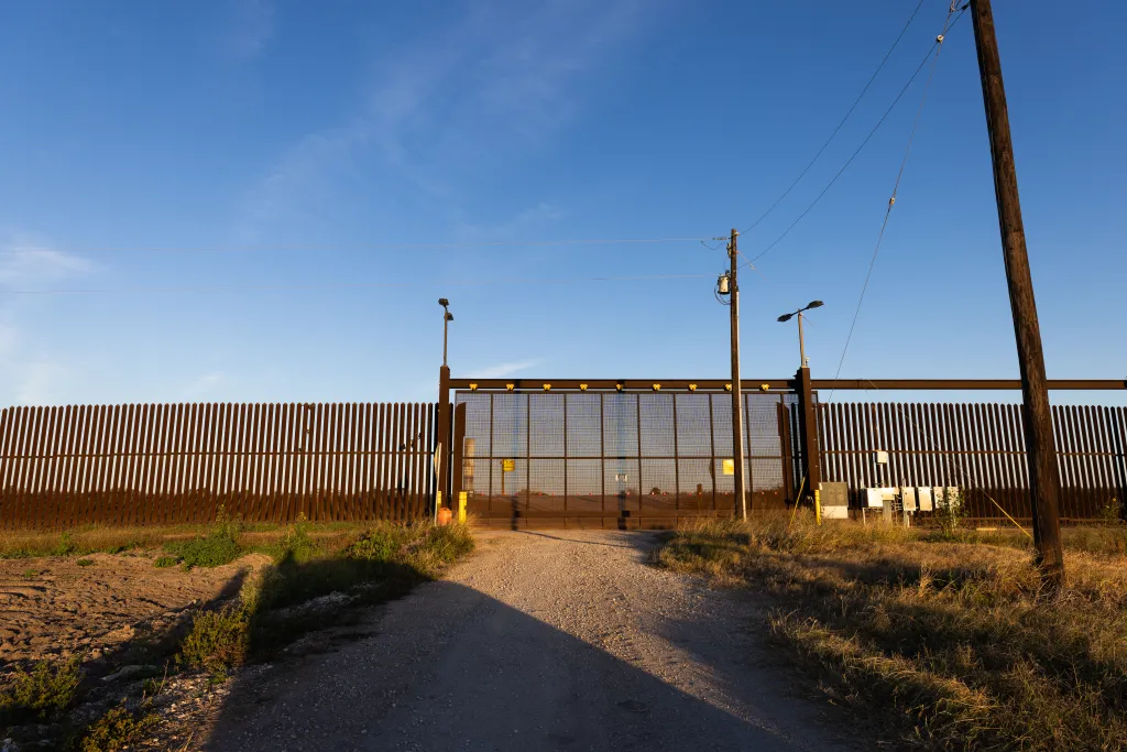 A U.S.-Mexico border wall with a large gate, power lines, and a dirt road in Brownsville, Texas.