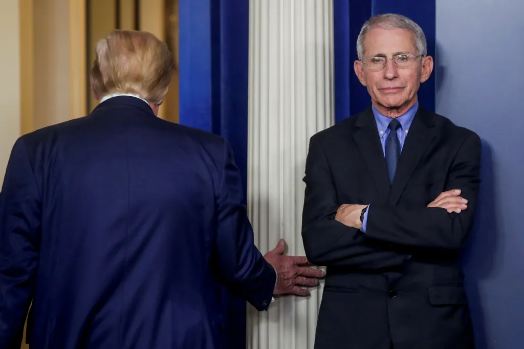 Dr. Anthony Fauci stands with his arms crossed while U.S. President Donald Trump, seen from behind, departs.