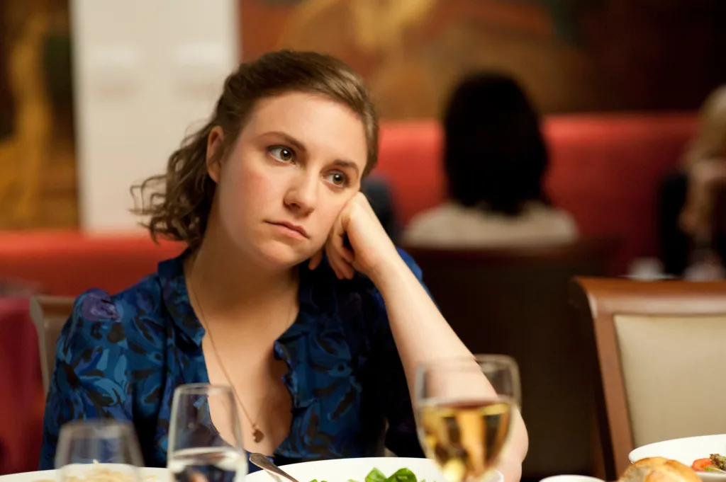 Lena Dunham resting her chin on her hand, looking bored at a restaurant table.