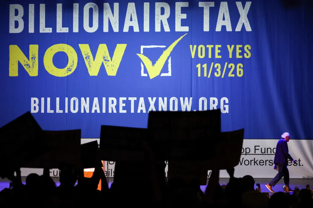 Senator Bernie Sanders walks past a "Billionaire Tax Now" banner with a checkbox and "Vote Yes 11/3/26" text.