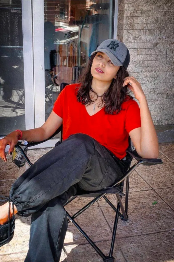 Venus Hosseininejad sitting in a folding chair, wearing a red shirt, black pants, and a New York Yankees baseball cap.