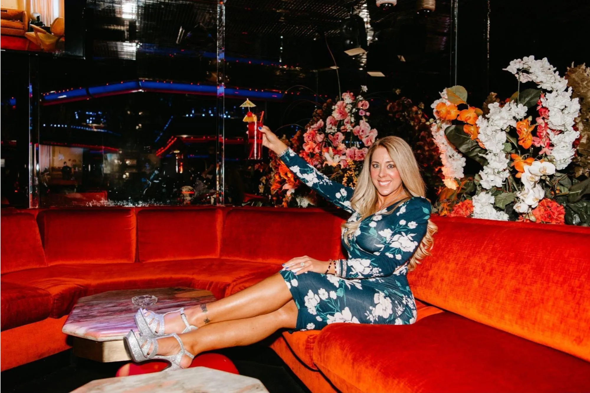 A woman in a floral dress holding a cocktail, sitting on a red velvet couch in a darkly lit room with floral decorations.
