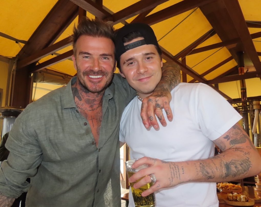 David and Brooklyn Beckham smiling, with Brooklyn holding a glass of beer.