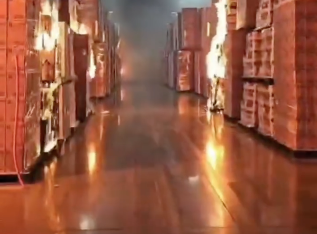 A warehouse aisle with stacked pallets of boxes, engulfed in flames and smoke.