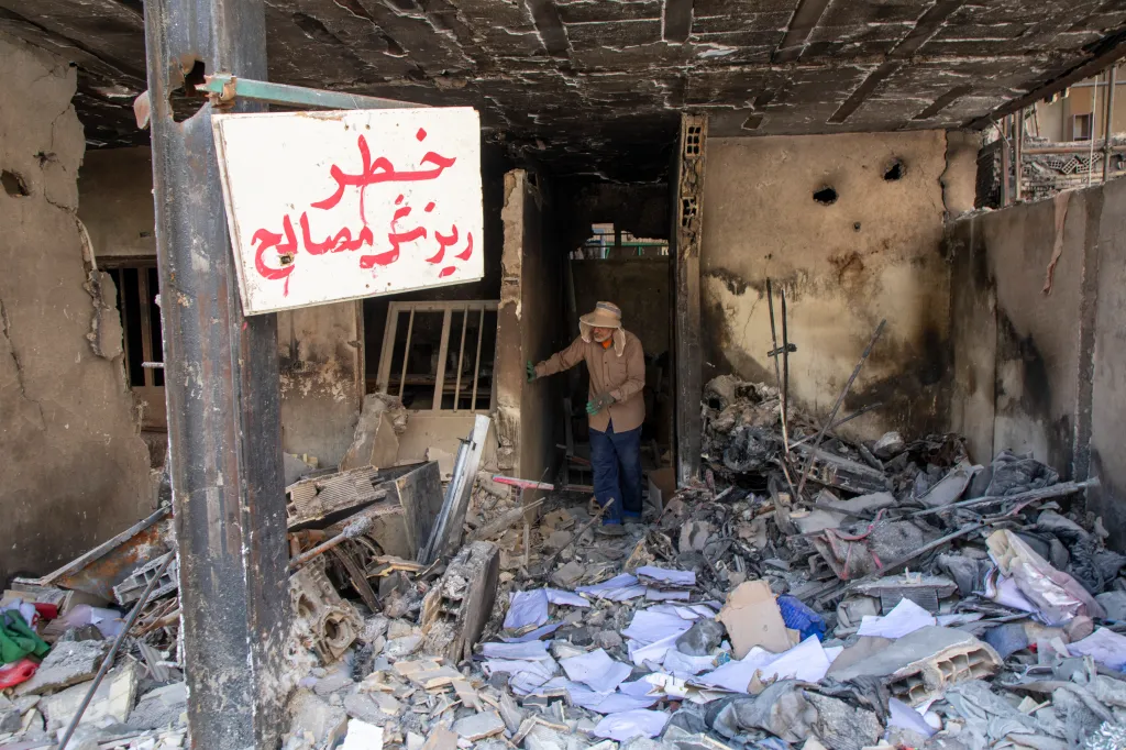 Man in a hat and gloves inspecting damage to a residential building in Tehran, Iran, with debris and a sign with red Arabic writing visible.