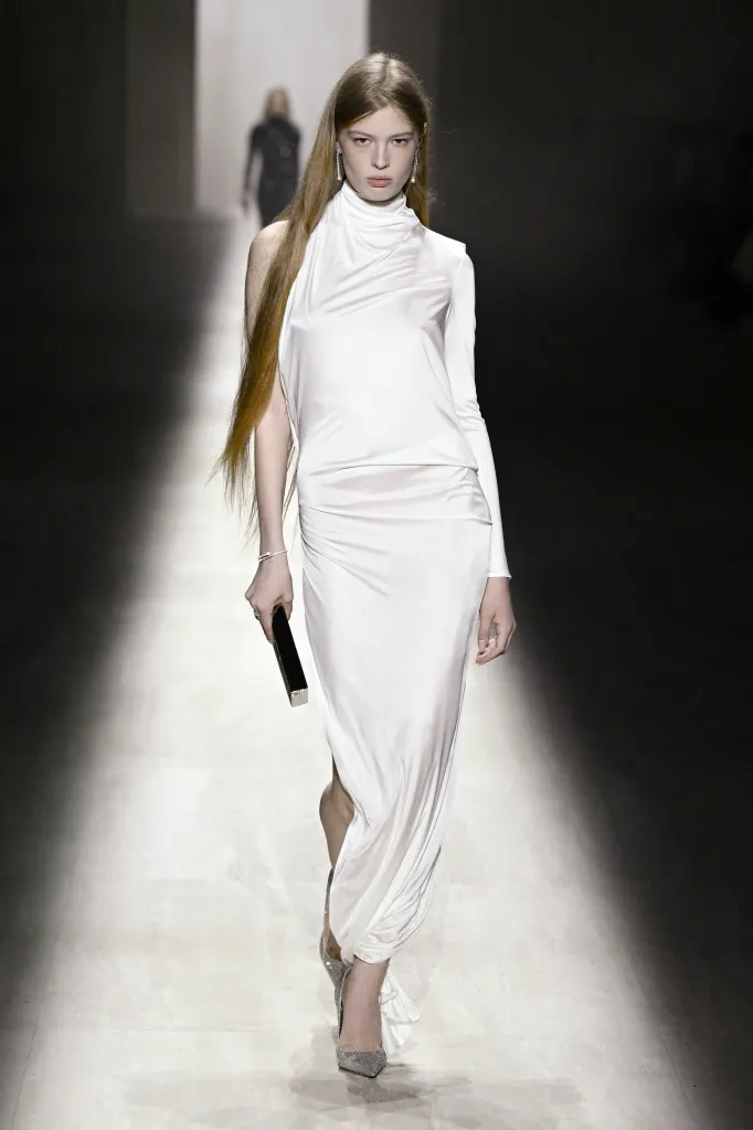 Vivian Jenna Wilson on the runway at the Gucci fashion show, wearing a long white gown and carrying a black clutch.