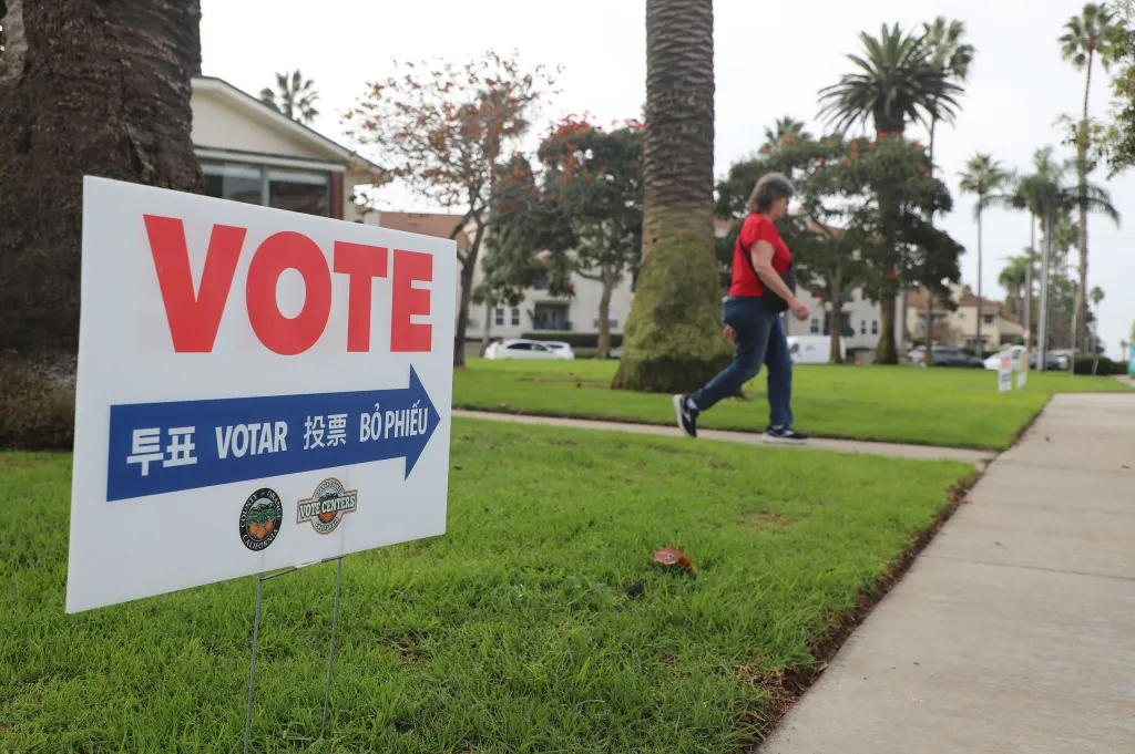 A "VOTE" sign in English, Korean, Spanish, Chinese, and Vietnamese with an arrow pointing right on a grassy lawn with a voter walking by.