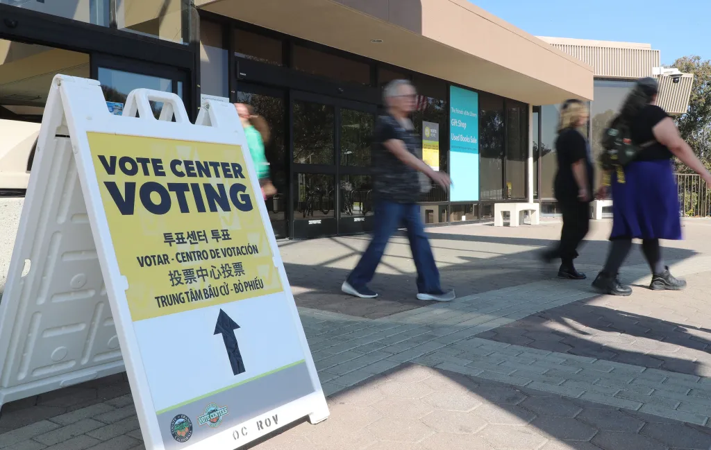 A "Vote Center Voting" sign with multi-lingual text and an arrow points towards the entrance of the Huntington Beach Central Library as blurred people walk past.